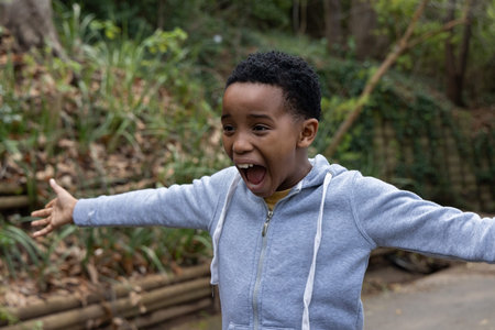 African American schoolboy standing on paved trail in gray hoodie, yellow shirt, shouting arms wide. Child, lifestyle, candid, outdoors, park, path, woodland