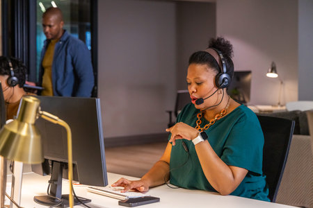 Diverse coworkers with woman in green top speaking into headset while typing at desk with monitor. Office, teamwork, openplan, headphones, microphone, smartwatch, callcenter