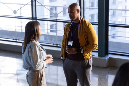 Diverse coworkers chatting in atrium by floor-to-ceiling windows holding coffee cups with lanyard. Colleagues, discussion, casual, lobby, daylight, denim, jacket