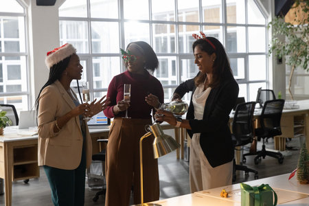 Diverse female team in businesswear wearing holiday hats pouring from bottle into flutes at office. Celebration, collaboration, professional, bright, openplan, glassware, decor