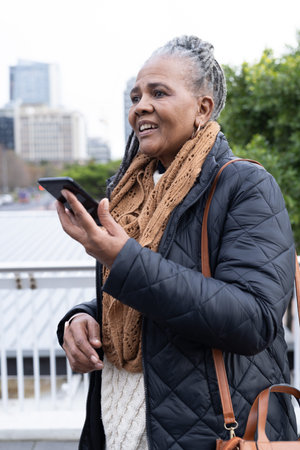 Vertical video: African American Senior woman on bridge, using phone assistant, holding brown bag. Elderly, smartphone, urban, outdoor, promenade, railing, scarf