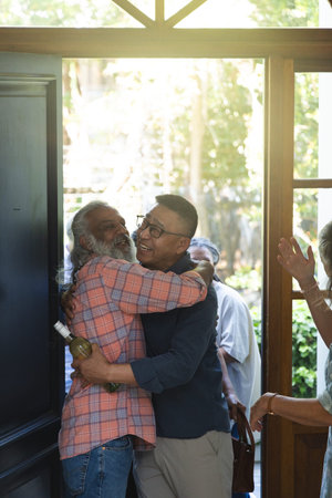 Diverse friends greeting, hugging at wooden door, bearded Indian man in plaid shirt holding wine. Threshold, sunlit, garden, foyer, hospitality, reunion, candid