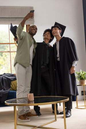 Diverse family wearing graduation gowns and mortarboards taking smartphone selfie in living room. Cap, men, home, sunlit, candid, portrait, celebration