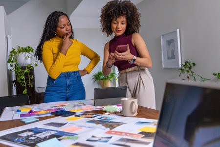 African American women in mustard, burgundy tops reviewing prints at wood table holding phone. Collaboration, brainstorming, moodboard, planning, design, studio, workspace