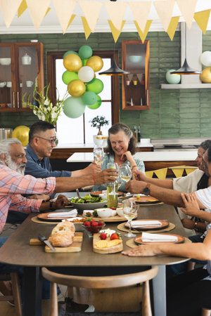 Host raising glass initiating toast, group clinking glasses, passing bread and sharing food. Gathering, festive, sunlit, dining, wooden, balloons, banners
