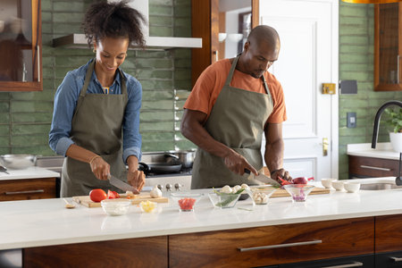 Couple chopping vegetables at countertop island wearing green aprons with wooden cutting boards. Duo, homemaking, kitchen, knives, mushrooms, tomato, onion