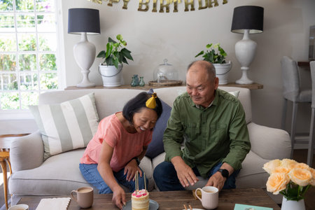 Asian couple with senior woman leaning and sitting on beige sofa celebrating with lit cake, roses. Cozy, living, decor, candles, banner, neutral, light