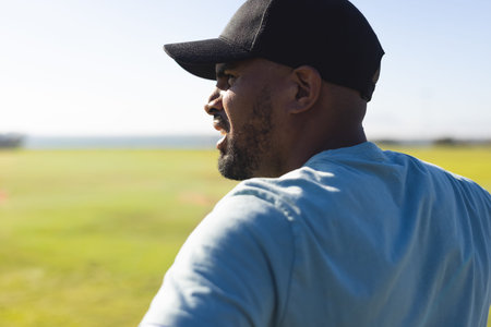 African American man standing in open field, calling, wearing black cap, light-blue tee, copy space. Outdoors, grassland, sunlit, profile, hat, casual, conversation