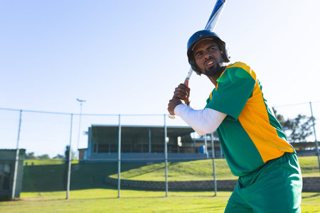 Asian man batting on baseball field wearing team uniform with helmet holding bat, copy space. Athlete, outdoor, grass, sunny, fence, bleachers, stadium