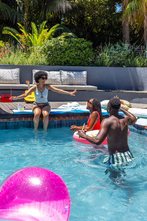 African American friends chatting and sipping at private backyard pool near pink inflatable ball. Group, outdoor, summer, tropical, deck, beverages, sunglasses