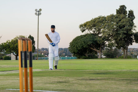 Man walking cricket pitch at dusk, white kit, pads, gloves, black cap, holding bat near stumps. Athlete, batter, field, twilight, floodlights, outdoor, sport