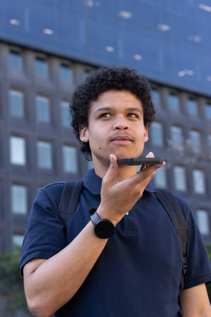 African American man speaking into smartphone by office tower in navy polo, backpack and smartwatch. Outdoor, urban, professional, commuter, technology, connectivity, corporate