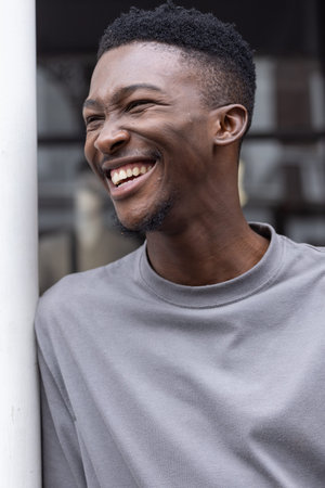 African adult male laughing, wearing light-gray crewneck, leaning against white pole by building. Portrait, smile, candid, urban, outdoor, lifestyle, diffused