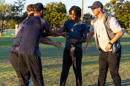 Diverse male teammates huddling on field in late sun, coach holding whistle, wearing track pants. Teamwork, outdoors, fitness, leadership, camaraderie, park, grass