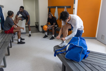 Diverse male teammates in locker room wearing gear, Asian man tying shoe near duffel, copy space. Basketball, gym, bench, headphones, teamwork, athleticwear, camaraderie