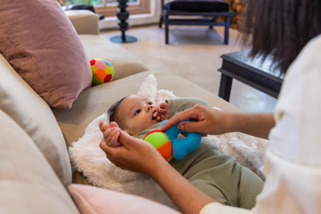 Female infant lying on white cushion on beige sofa at home, caregiver holding colorful ring toy. Baby, bonding, daylight, cozy, pastel, window, plush