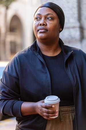 African American woman wearing black jacket standing on sidewalk holding cup near arch looking left. Headwrap, urban, street, coffee, archway, textured, diffused