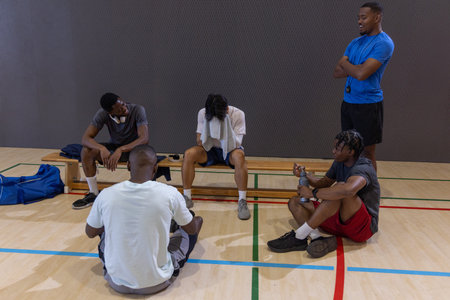 Diverse male teammates resting on wooden bench on gym court, holding water bottle and towel. Basketball, team, athletes, training, recovery, group, focus