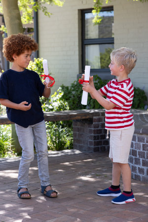 Diverse boys exchanging rolled white scrolls tied with red ribbons on paved brick patio near window. Children, outdoor, courtyard, sunlit, shadows, greenery, friendship
