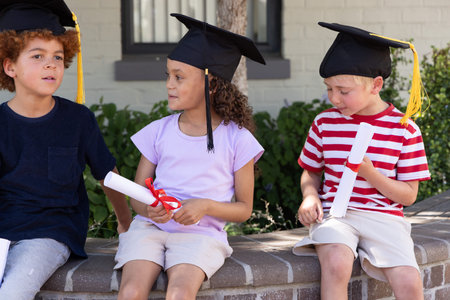 Diverse children sitting on curved brick bench at school wearing caps with tassels holding diplomas. Mortarboard, vibrant, outdoor, courtyard, ceremony, celebration, students