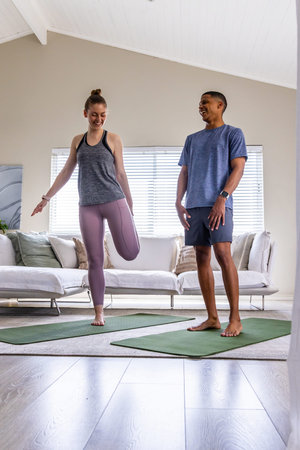 Diverse couple in activewear following man's cue, practicing balancing on green mats in living room. Yoga, balance, fitness, duo, home, daylight, interior