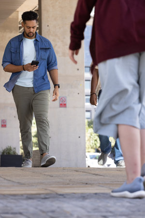 African American man walking toward lens while using smartphone under pillars with red signs. City, pedestrian, daylight, phone, street, concrete, commuter