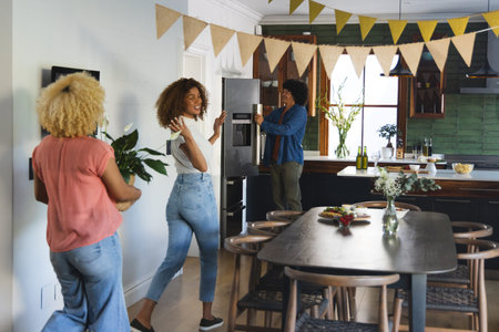 Three adult African American women decorating kitchen with bunting, holding potted plant by fridge. Gathering, friends, home, dining, decor, celebration, cozy