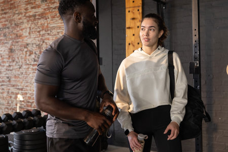 Male trainer and woman trainee chatting between sets near squat rack holding bottle shaker gym bag. Strength, conversation, partners, fitness, industrial, brick, weights