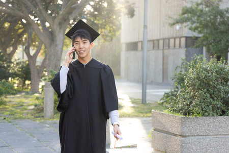 Asian man in black gown and mortarboard holding diploma, talking on smartphone walking campus path. Grad, cap, outdoor, backlit, university, lamppost, greenery