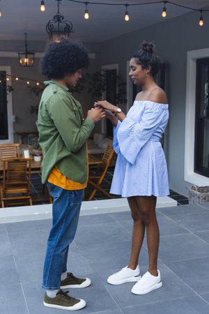 Couple, African American man in green jacket placing ring on woman in striped dress on patio