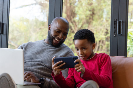 African American father and son sitting on couch in living room watching smartphone, laptop visible