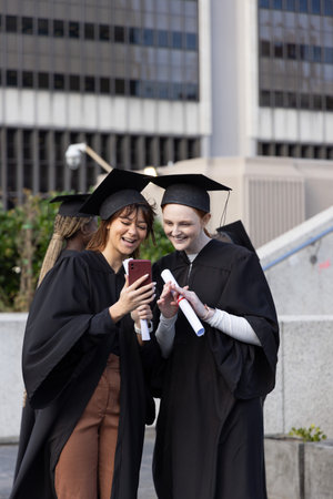 Female classmates looking at red phone, wearing mortarboards and holding diplomas on stone steps