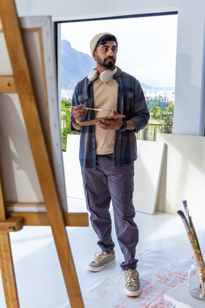Indian man standing in art studio wearing beanie and headphones holding palette and brush