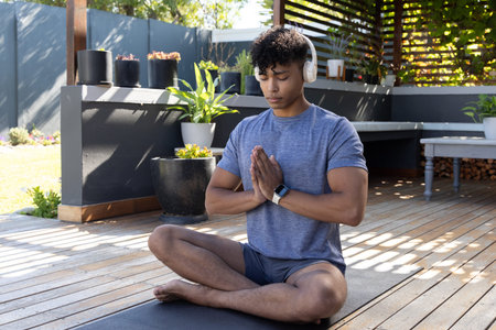 African American man meditating on yoga mat in yard wearing athletic wear, headset, smartwatch