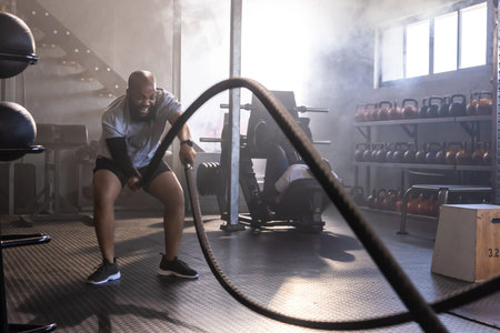 African American men swinging battle ropes and using weight machine in gritty gym, copy space