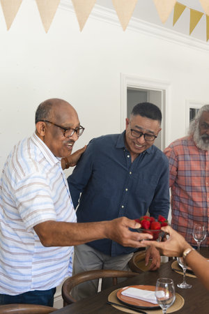Senior men smiling, reaching, passing bowl of strawberries around wooden dining table at home