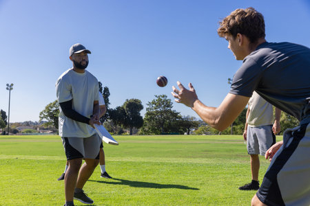 Male teammates practicing football drill on grassy field, catching brown football, holding towel