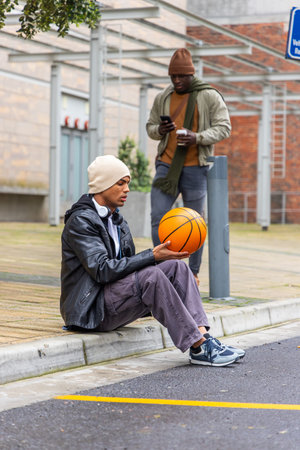 Diverse male pair sitting on curb at plaza holding orange basketball with headphones checking phone