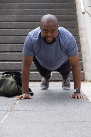 Adult African man doing push-ups on slabs at base of stairs, wearing heather tee, smartwatch