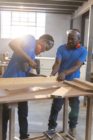 African American coworkers using power drill securing plank in workshop wearing goggles, earmuffs