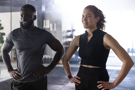 Female and male exercisers standing with hands on hips at gym, wearing fitted gear and smartwatches