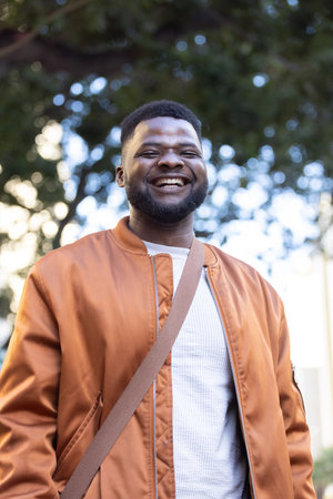 Adult man standing smiling in park, wearing rust bomber jacket, white textured sweater, tan strap