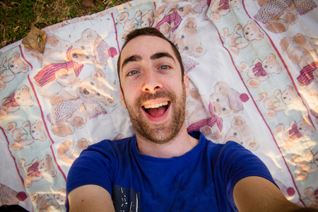 Young man lying on a sheet in the park and taking selfie picture with goofy smile.の写真素材