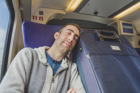 Young man traveling on a train and sleep on blue suitcase next to him.の写真素材