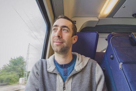 Young man traveling on a train and looks out the window and smiling. A blue suitcase is next to him.