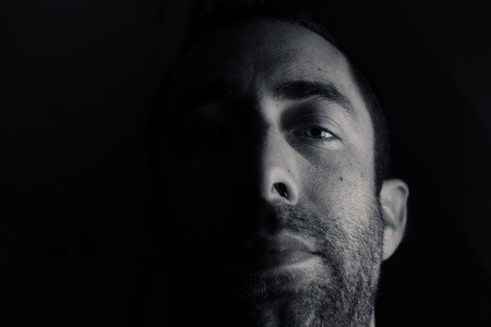 Dramatic portrait of a young man half expressionless face in deep shadow looking at camera isolated...