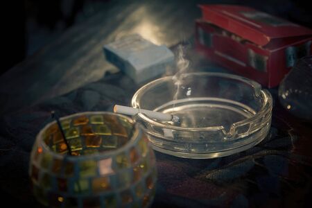 Smoke emitted from a burnt cigarette in a glass ashtray. It placed on a tablecloth on a wooden table. Close up, loop with selective focus.の写真素材