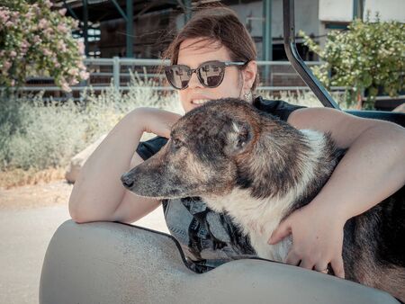 Caucasian woman is holding her dog while traveling. They riding in an electric golf cart in the nature of northern Israel on summer sunny day. Mixed breed dog with cropped ears.の写真素材