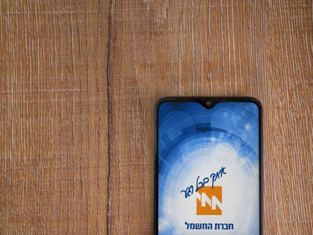 Lod, Israel - July 8, 2020: Israel Electric Corporation app launch screen with logo in hebrew text on the display of a black mobile smartphone on wooden background. Top view flat lay with copy space.のeditorial素材