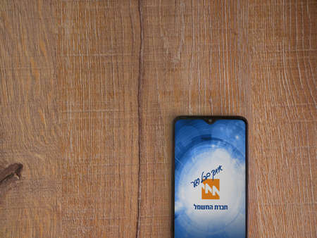 Lod, Israel - July 8, 2020: Israel Electric Corporation app launch screen with logo in hebrew text on the display of a black mobile smartphone on wooden background. Top view flat lay with copy space.のeditorial素材